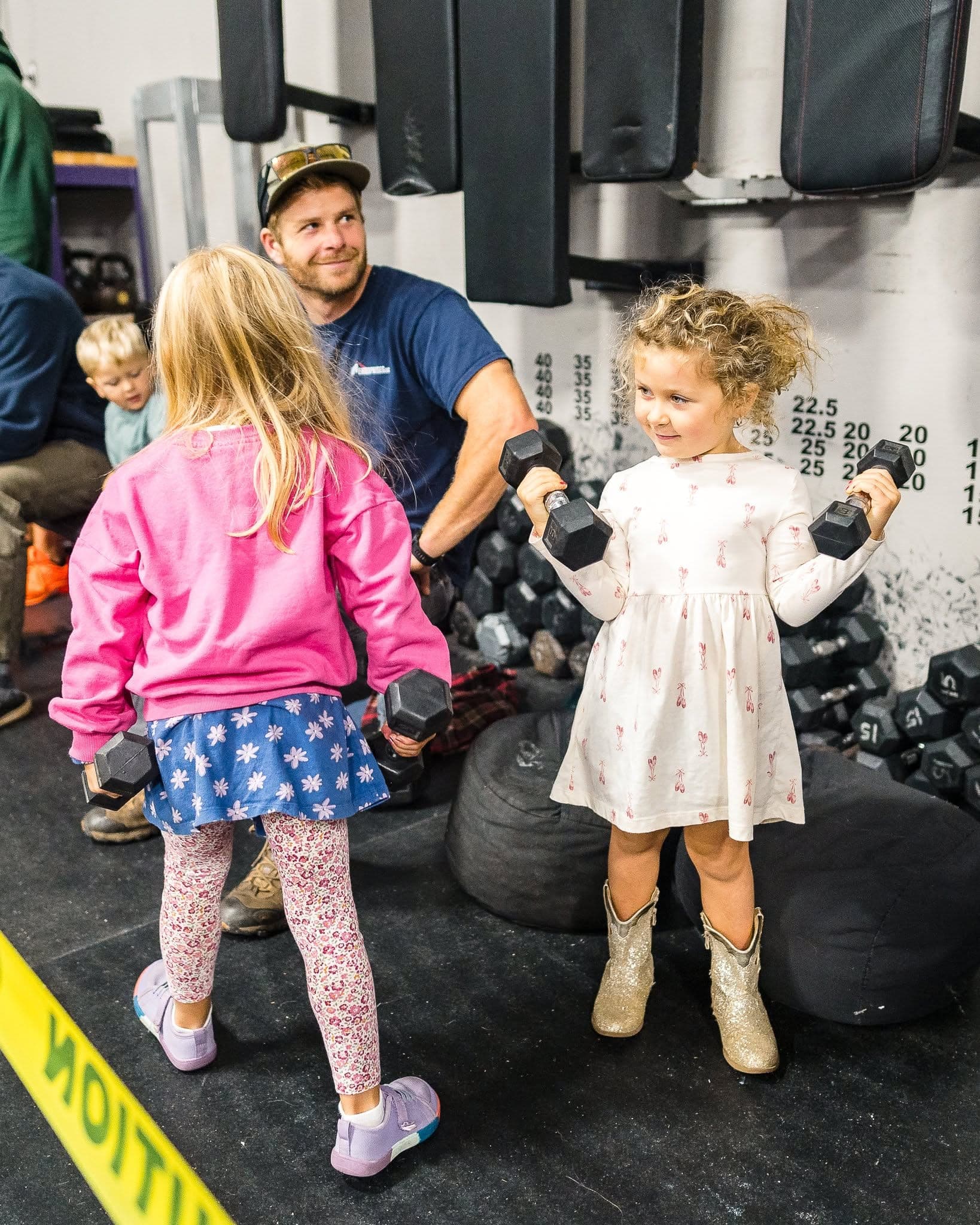 A coach in a cap smiling with three young children holding small dumbbells on the gym floor